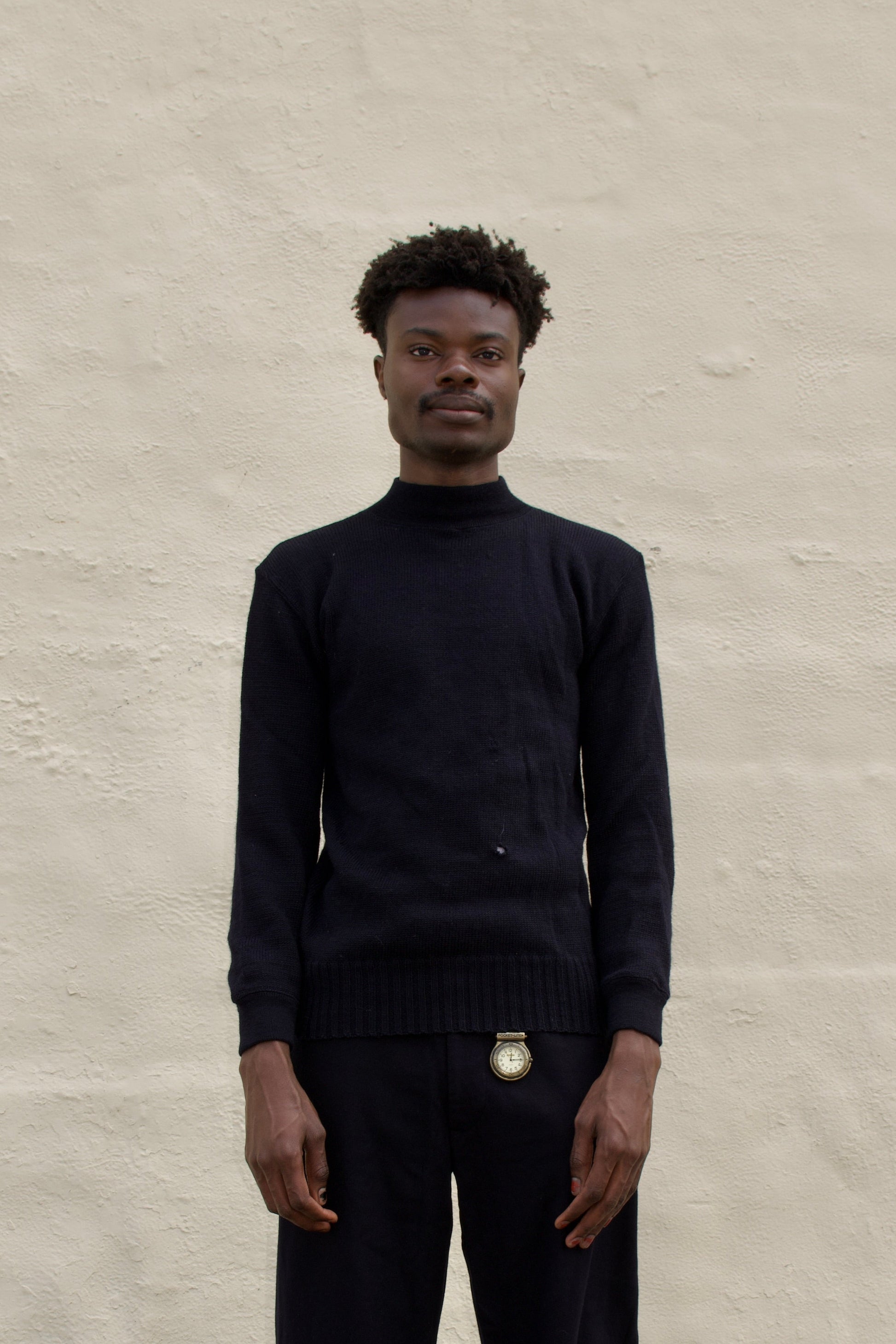 Person wearing a black turtleneck and pants standing against a plain wall.