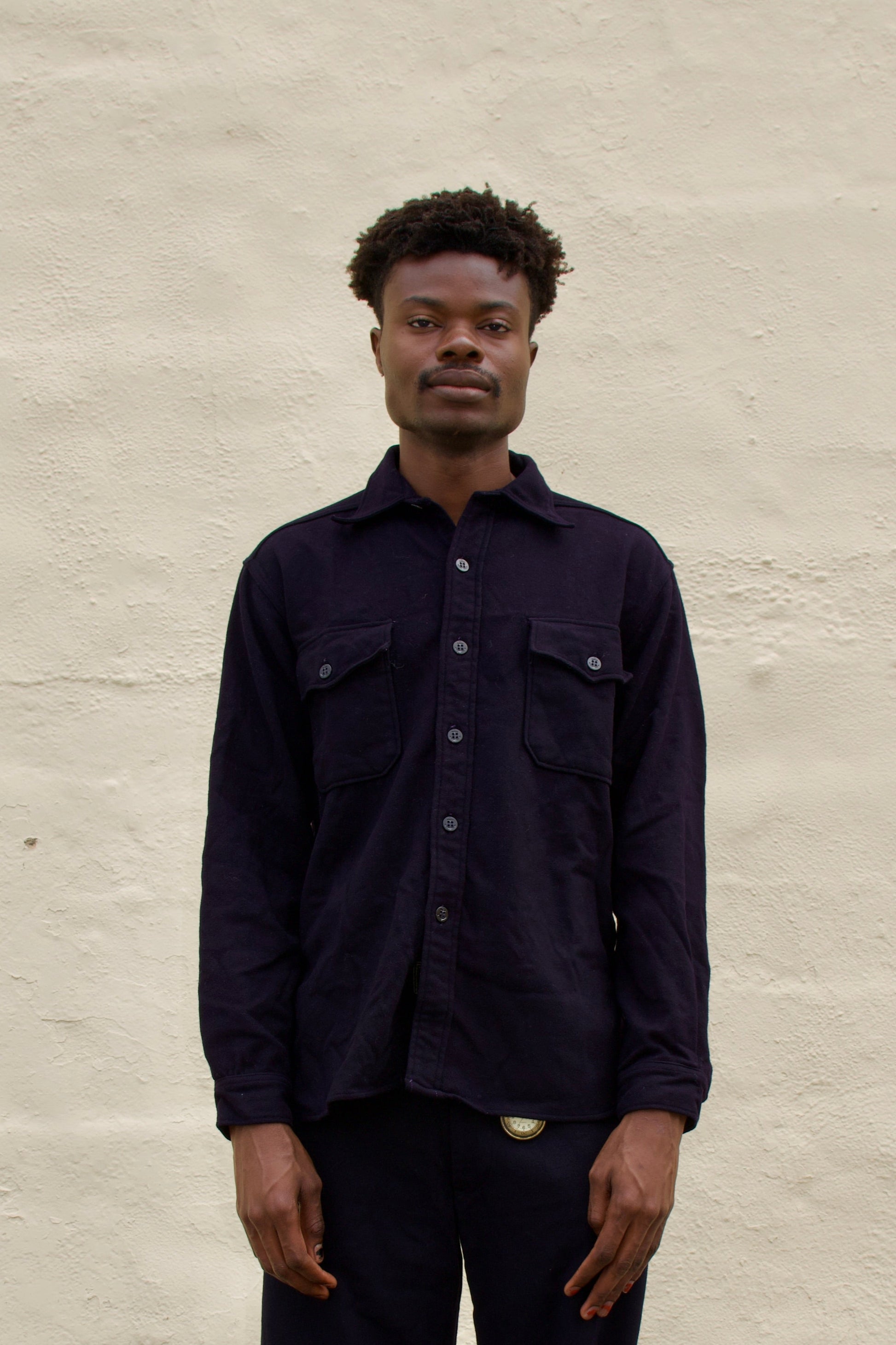 Man wearing a dark shirt and pants standing against a light beige wall.