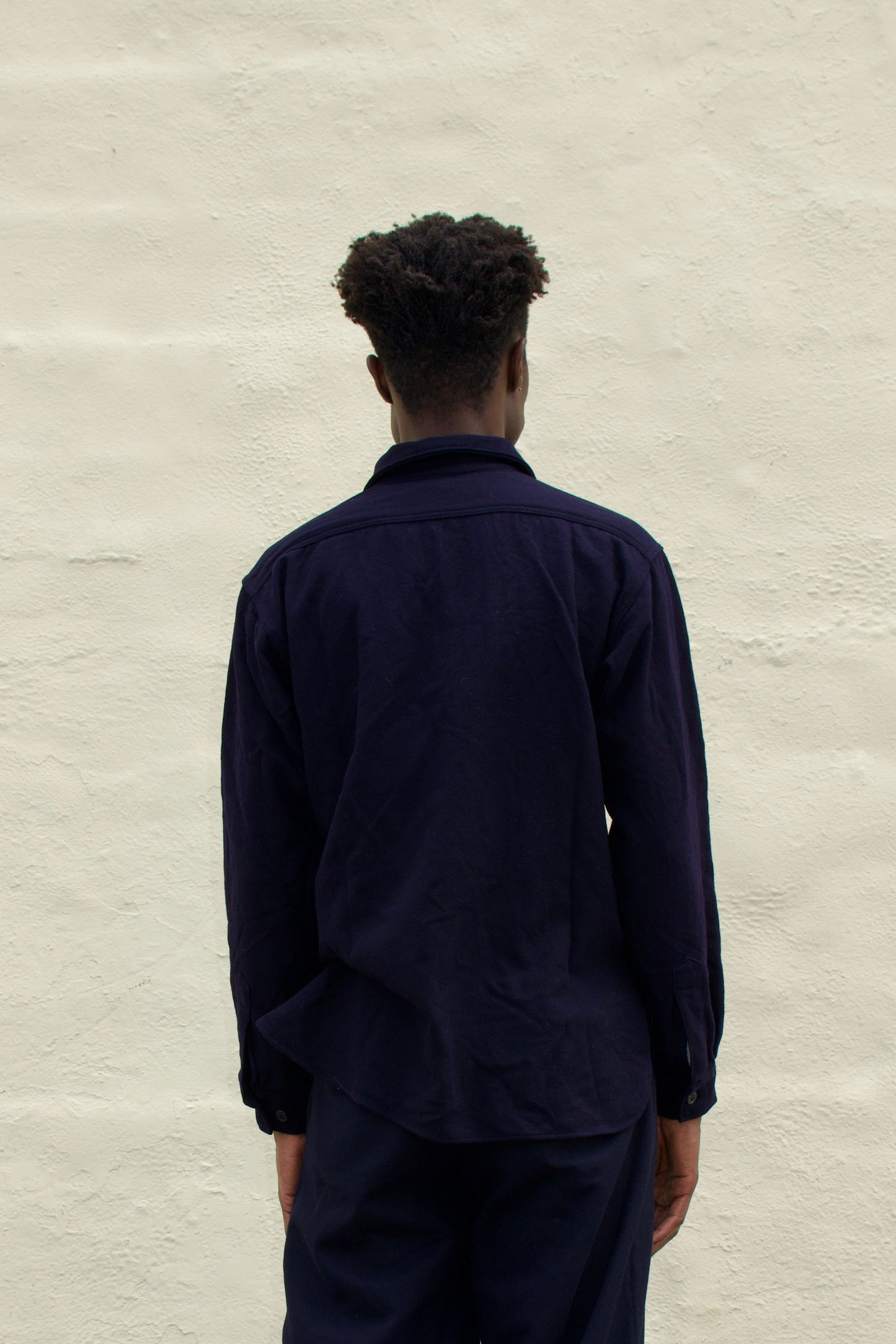 Person wearing a dark outfit standing against a light gray wall.