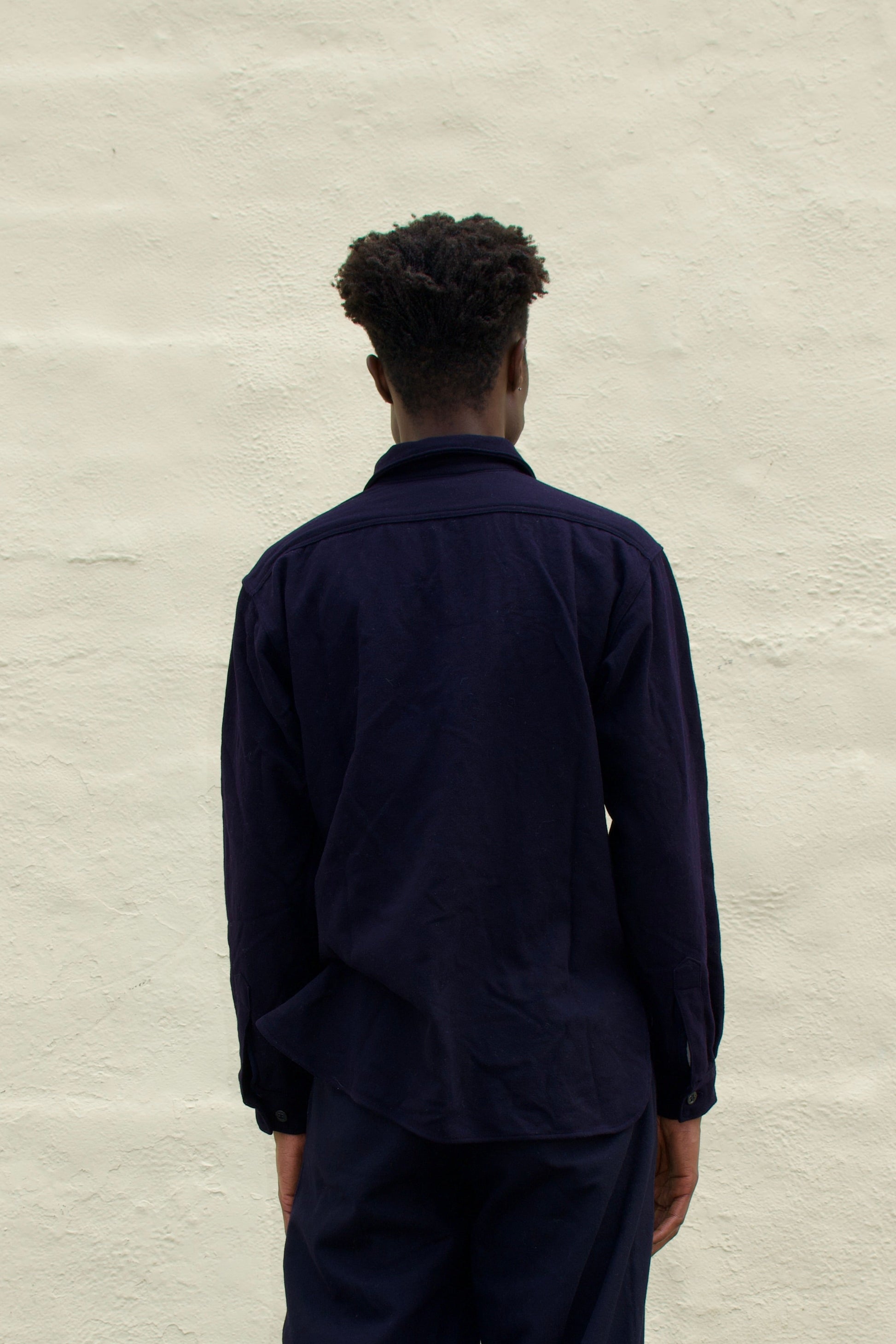 Person wearing a dark outfit standing against a light gray wall.